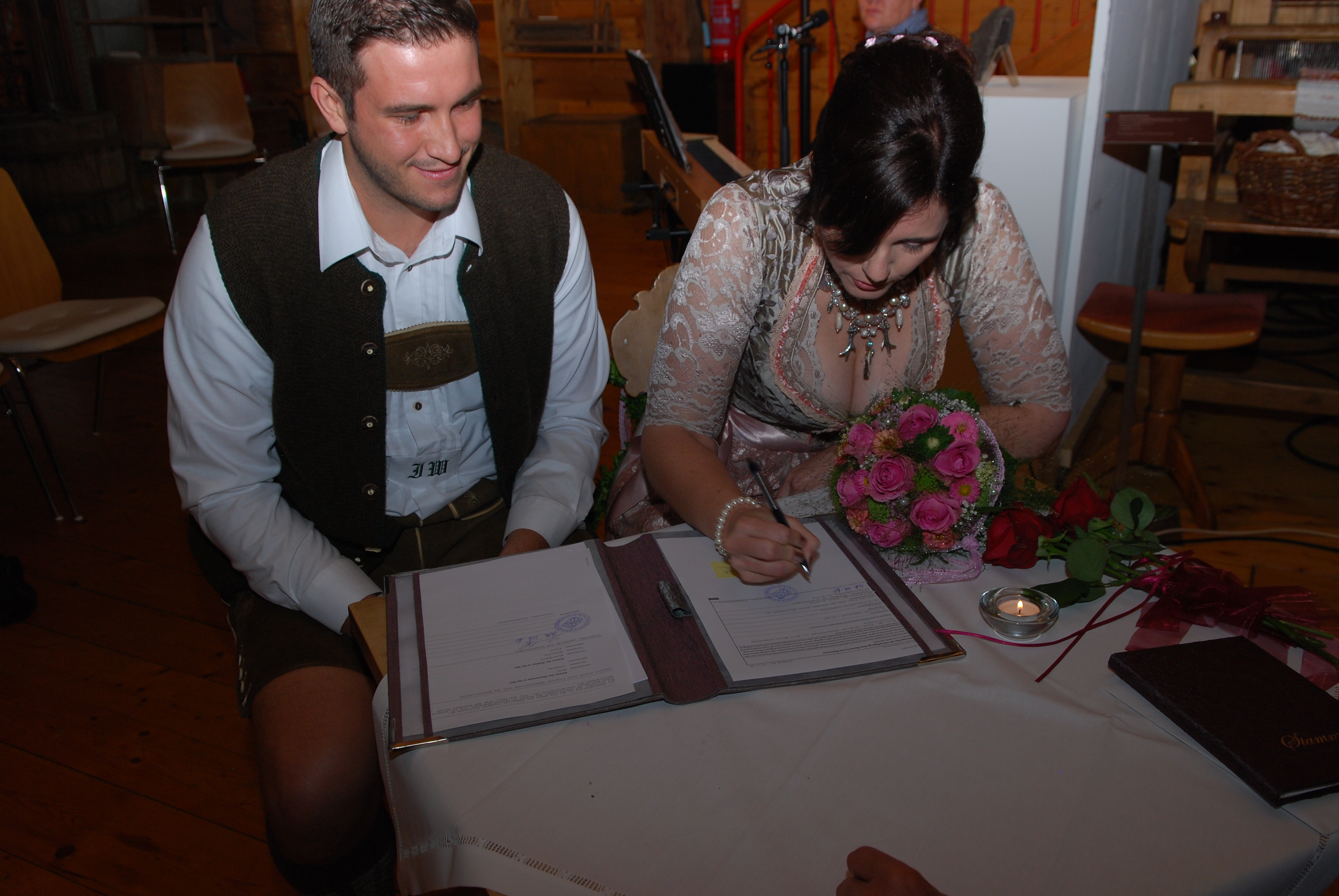 Brautpaar in Tracht sitzt an einem geschmückten Tisch, die Frau setzt ihre entscheidende Unterschrift während der Mann lächelnd zusieht - das Bild wird mit Klick vergrößert 