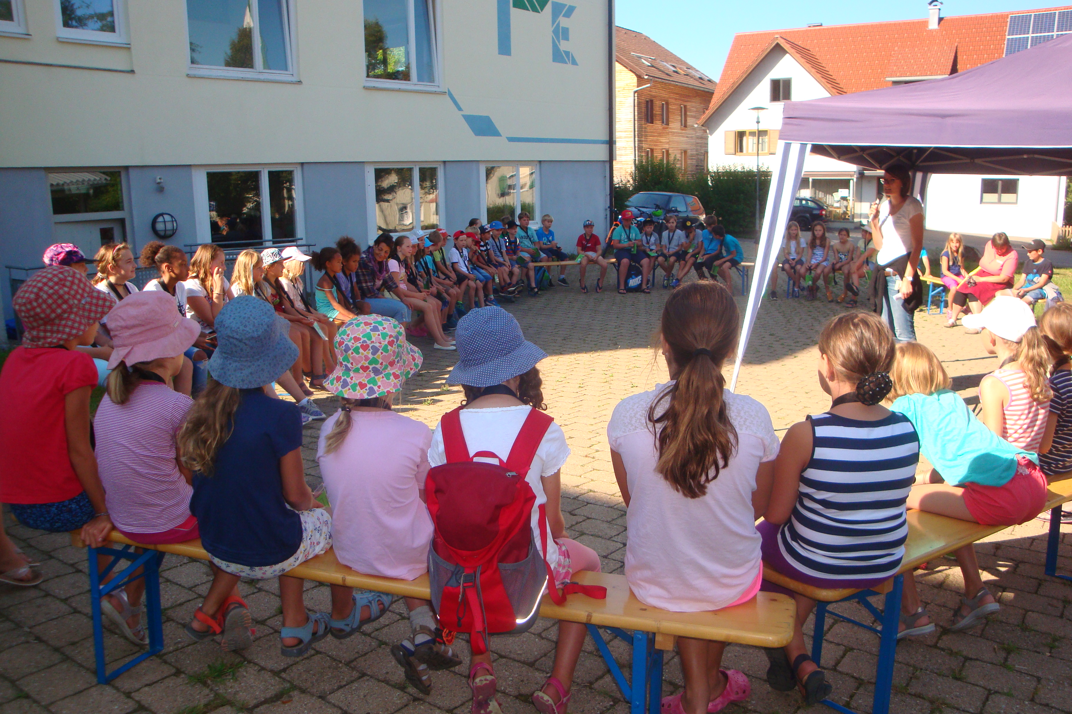  Kinder sitzen auf Bänken in einem großen Kreis, in der Mitte ist ein Pavillon aufgebaut - das Bild wird mit Klick vergrößert 