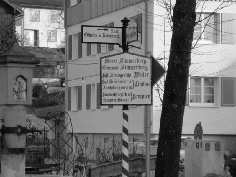  Altes Ortsschild Simmerberg mit Haus im Hintergrund - Bild in schwarzweiß - das Bild wird mit Klick vergrößert 