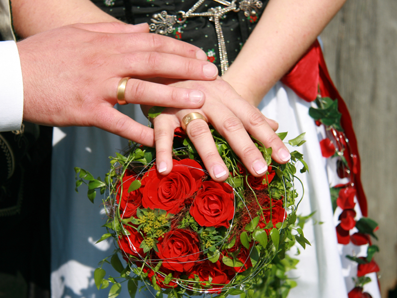  Ehepaar hält jeweils die Hand mit dem goldenen Ehering über einen Strauß Blumen aus roten Rosen - das Bild wird mit Klick vergrößert 